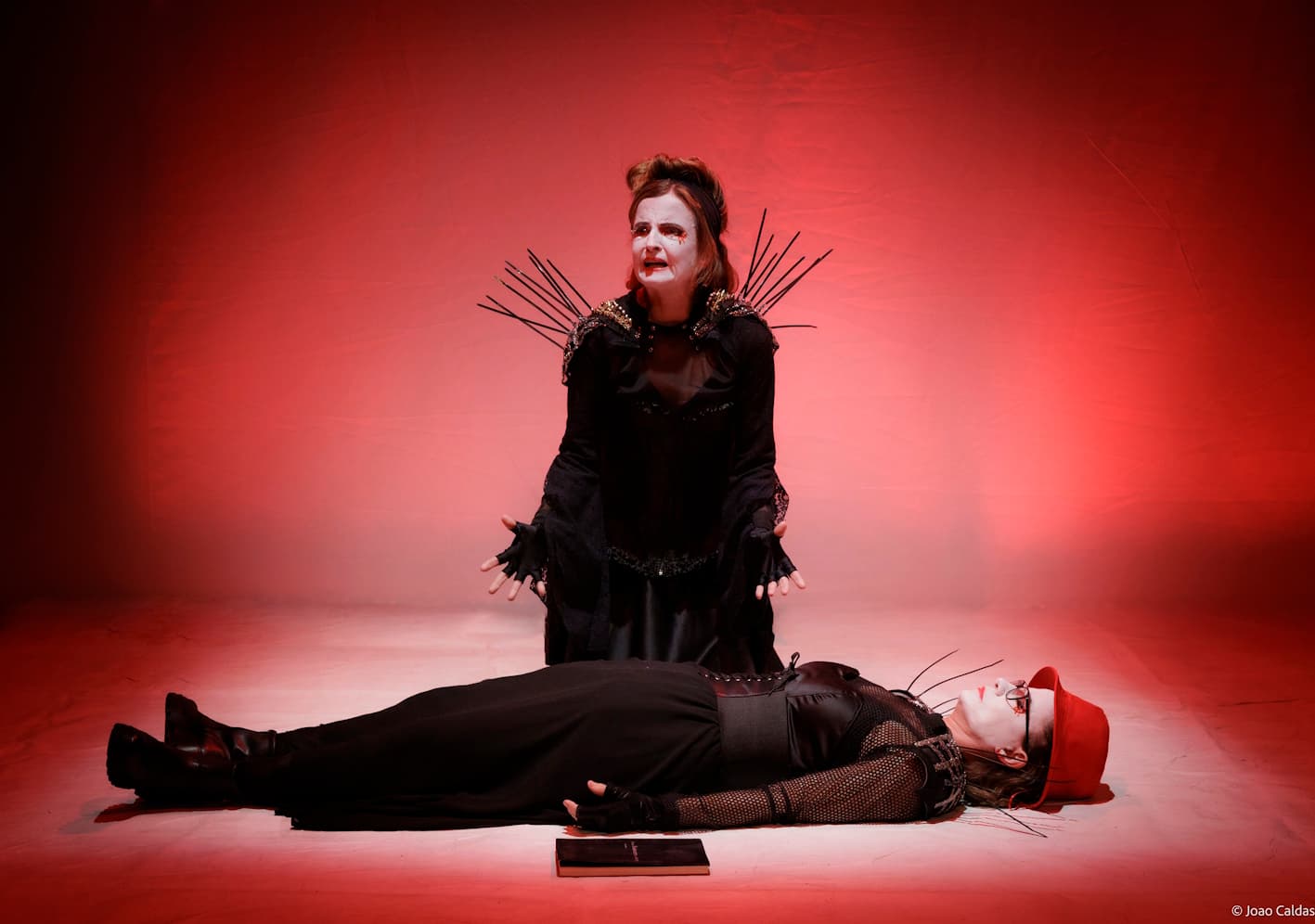 Tableau em luz vermelha: uma intérprete ajoelhada em gesto ritual, outra deitada; atmosfera trágica.